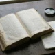 Open Latin dictionary beside a notebook and magnifying glass on a wooden desk.